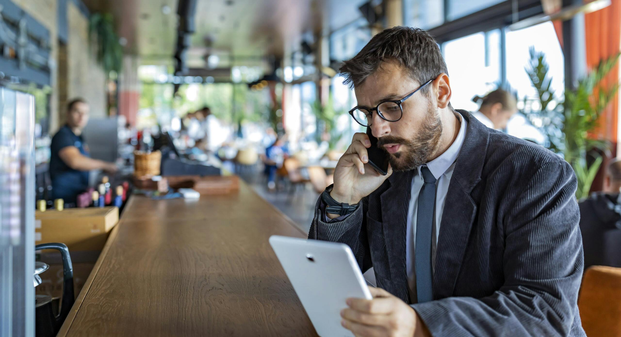 Happy and Successful Male Business Person with Beard, Dressed in Casual Suit is Communicating over a Mobile Phone and Drinking Coffee in Modern Restaurant. A Modern and Handsome Businessman is Sitting in City Café, Enjoying in Hot Cappuccino and Talking about Business with his Colleagues.